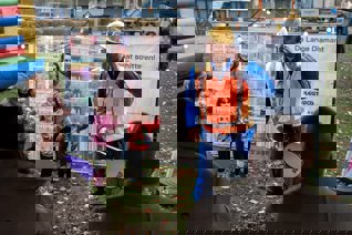 3 kleine kinderen staan met hun moeders bij een bijzonder bouwhekdoek met hun kleurtekeningen en artist impressie met een bouwvakker met een trommel