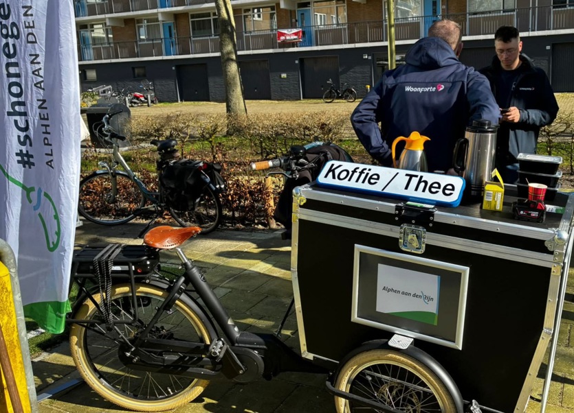 De fietskar met koffie en thee staat voor de flat Plutostraat met wijkbeheerder van Woonforte op de rug gezien met logo Woonforte op de blauwe jas
